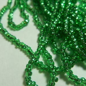 May include: A strand of green seed beads with a metallic sheen. The beads are small and round, and they are strung together in a long strand.