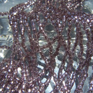 May include: A close-up of a strand of light purple glass seed beads. The beads are small and round, and they have a slight iridescent sheen.