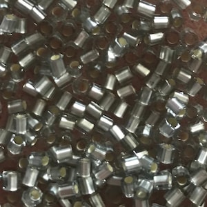 May include: A close-up image of a large quantity of silver-colored glass beads. The beads are small and cylindrical in shape. They are scattered on a brown surface.