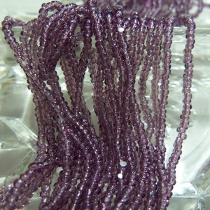 May include: A close-up of a strand of small, round, purple glass beads. The beads are arranged in a loose pile, and the light reflects off of their surfaces.