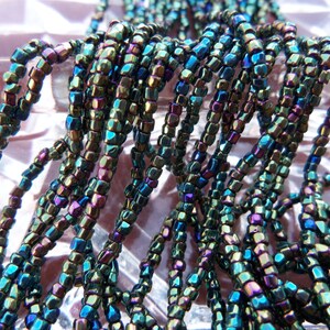 May include: Close-up of strands of iridescent beads. The beads are small, square-shaped, and have a metallic sheen with colors ranging from teal and blue to purple and bronze. The beads are strung together, creating a textured and layered effect.