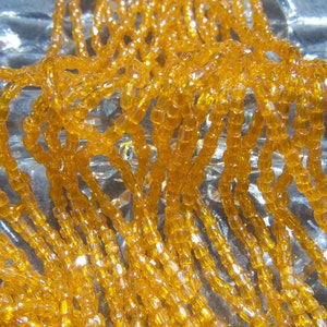 May include: A close-up of a strand of amber-colored glass beads. The beads are small and round, and they are strung together on a clear thread. The beads are arranged in a random pattern, and they create a shimmering effect.