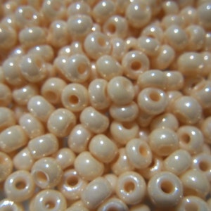 May include: A close-up of a large quantity of small, round, off-white beads with a pearlescent finish. The beads have a small hole in the center for stringing.
