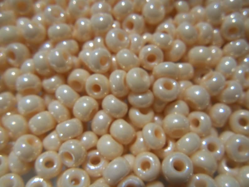 May include: A close-up of a large quantity of small, round, off-white beads with a pearlescent finish. The beads have a small hole in the center for stringing.