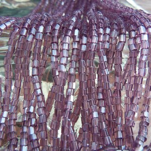 May include: A strand of purple glass seed beads. The beads are square shaped and have a slight iridescent sheen.