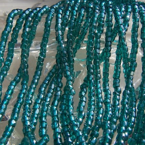 May include: A close-up of a strand of teal green glass seed beads. The beads are small and round, and they have a slight iridescent sheen.