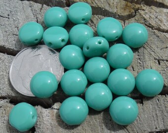 May include: A pile of turquoise blue glass beads, each with two holes. The beads are round and smooth, and they are scattered on a wooden surface.