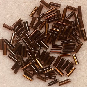 May include: A large quantity of small, dark brown glass beads with a gold iridescent sheen. The beads are cylindrical in shape and are scattered on a white surface.
