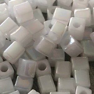 May include: A close-up of a pile of white iridescent square beads. The beads are small and have a hole in the center.