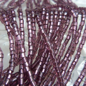 May include: A close-up of a strand of purple glass beads. The beads are small and have a square shape. They are arranged in a random pattern.