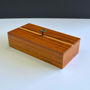 May include: A rectangular wooden box with a warm brown finish and a light-colored stripe down the center of the lid. The box has a small, decorative knob. The box is likely used for storage or display.