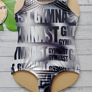 May include: A sleeveless leotard with the word "GYMNAST" in large, bold, black letters repeated across the front. The leotard has a gradient design of black, white, and grey. The leotard is displayed on a wooden form.
