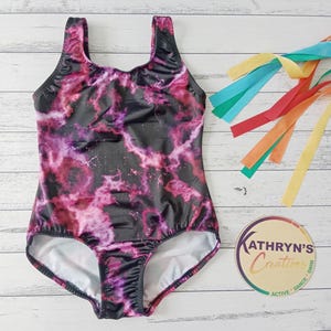 May include: A black and pink galaxy-print leotard with a scoop neck and a high-cut leg. The leotard is designed for active wear and dance. The image also includes colourful ribbons and a logo that says "Kathryn's Creations".