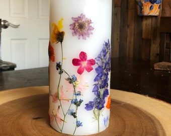 Può includere: Una candela bianca a forma di pilastro con fiori pressati incorporati nella cera. I fiori sono in tonalità di rosa, viola, giallo, arancione e blu.