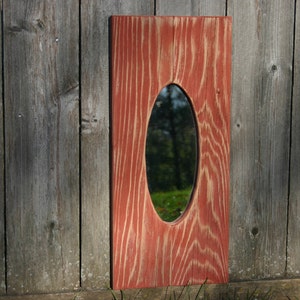 May include: A rustic wooden mirror with a red stain finish and an oval-shaped mirror inset. The mirror is hanging on a wooden wall.