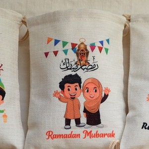 May include: Two children, a boy and a girl, are smiling and waving. They are dressed in traditional clothing and are standing in front of a white bag with a red banner that says "Ramadan Mubarak". The bag is decorated with colorful flags and a lantern.