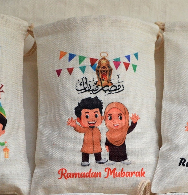 May include: Two children, a boy and a girl, are smiling and waving. They are dressed in traditional clothing and are standing in front of a white bag with a red banner that says "Ramadan Mubarak". The bag is decorated with colorful flags and a lantern.