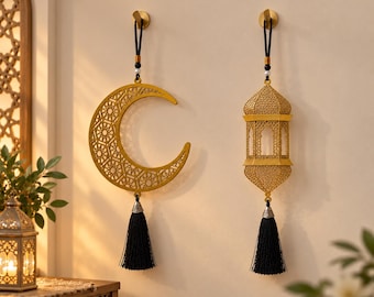 May include: Two gold-colored hanging ornaments featuring intricate geometric cutouts. One is a crescent moon and the other is a lantern shape. Both pieces are suspended by black cords with small beads and finished with long black fabric tassels.