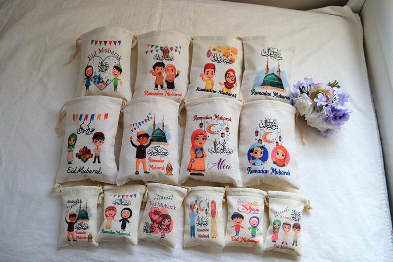 May include: A collection of 12 small, white fabric drawstring bags with colorful printed designs featuring cartoon characters, Islamic calligraphy, and the phrase "Eid Mubarak" or "Ramadan Mubarak".