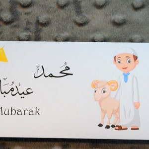 May include: White envelope with a cartoon illustration of a boy holding a lamb. The envelope has the text "Eid Mubarak" in English and Arabic. A $20 bill is sticking out of the envelope.