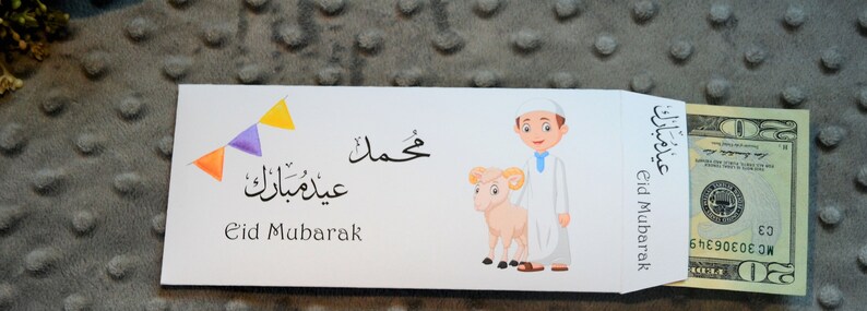 May include: White envelope with a cartoon illustration of a boy holding a lamb. The envelope has the text "Eid Mubarak" in English and Arabic. A $20 bill is sticking out of the envelope.