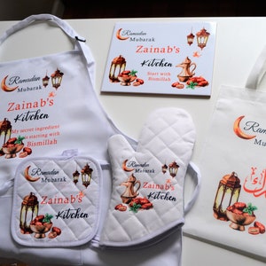 May include: A white kitchen apron, oven mitt, and tote bag with a Ramadan Mubarak theme. The items feature a crescent moon, lanterns, and the text "Zainab's Kitchen".