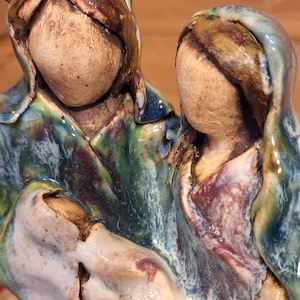 Può includere: Una scultura in ceramica raffigurante una famiglia, con figure in abiti con cappuccio. L'opera presenta una tavolozza di colori terrosi, tra cui marrone, verde e blu, con finitura lucida. Le figure sono vicine, suggerendo un tema di unione.