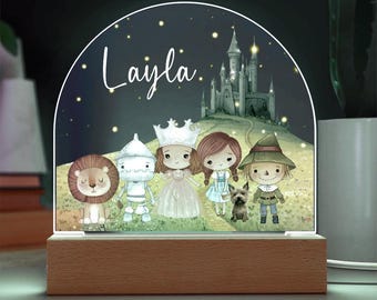 May include: An arched acrylic night light on a rectangular wooden base featuring a colorful illustration of a lion, a tin person, a princess, a girl, a small dog, and a scarecrow standing before a castle with the name Layla printed.