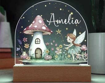 May include: An arched acrylic night light with a wooden base featuring a printed illustration of a mushroom house, a fairy, and a small frog. The name Amelia is printed in white script. The scene includes colorful flowers and glowing fireflies.