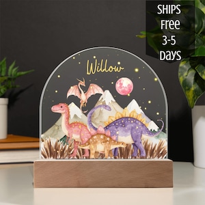 May include: A semi-circular acrylic night light featuring a watercolor illustration of a pink dinosaur, a purple dinosaur, a tan dinosaur, and a flying dragon. The scene includes mountains, a pink moon, and the name Willow in gold script.