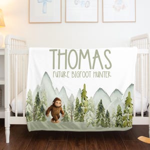 May include: A white baby blanket with the name "THOMAS" and the text "FUTURE BIGFOOT HUNTER" in green lettering. The blanket features a watercolor design of mountains, trees, and a brown Bigfoot figure. The blanket is draped over a white crib.