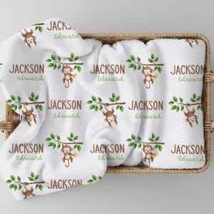 May include: White baby blanket with a repeating pattern of brown monkeys hanging from green branches, with the name "JACKSON Edward" in brown and green. The blanket is folded in a wicker basket.