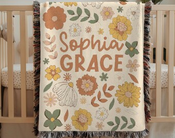 May include: A cream-colored baby blanket with a floral pattern and the names "Sophia Grace" in large, orange letters. The blanket features various flowers in shades of orange, yellow, and green, with fringe along the edges. The blanket is draped over a wooden crib.