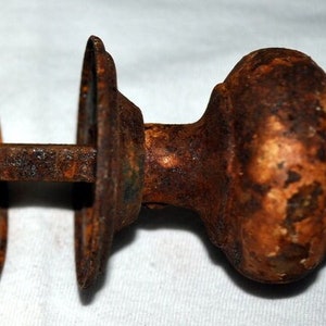 May include: A pair of antique door knobs, heavily rusted with a mottled orange and brown patina. The knobs have a rounded, floral-like design and are connected by a short, square rod. The backplates are circular with a decorative edge.