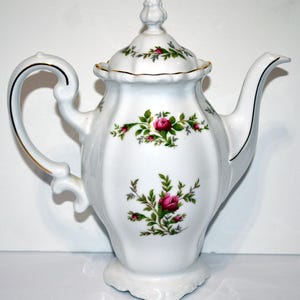 May include: White porcelain teapot with a lid, decorated with pink roses and green leaves. The teapot has a curved handle and spout, with gold trim accents. The base is a scalloped design, and the lid has a small finial.