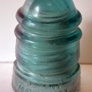 May include: A vintage teal glass insulator with a rounded top and a ribbed body. The base of the insulator has the text "PATENT MAY 2 1893" embossed on it. The glass has a slight iridescence.