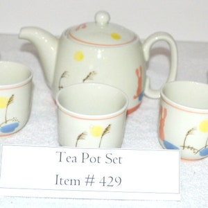 May include: A white tea pot set with a whimsical design featuring a yellow sun, blue water, and brown reeds. The tea pot and cups are decorated with a simple, hand-painted style. The set includes a teapot and four cups.  Tea Pot Set Item # 429