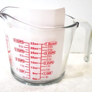 May include: A clear glass measuring cup with red markings for ounces and cups. The markings include 4 cups, 32 ounces, 1 quart, 3 1/2 cups, 28 ounces, 3 1/3 cups, 3 cups, 24 ounces, 3 cups, 2 1/2 cups, 20 ounces, 2 2/3 cups, 2 cups, 16 ounces, 2 cups, 1 1/2 cups, 12 ounces, 1 2/3 cups, 1 cup, 8 ounces, 1 cup, 1/2 cup, 4 ounces, 2/3 cup.