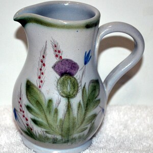 May include: A ceramic pitcher with a light blue and white base color. The pitcher features a hand-painted design of a purple thistle flower, green leaves, and red and blue accents. The handle is curved and the rim is slightly flared.