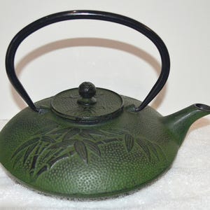 May include: A dark green cast iron teapot with a textured, pebbled surface and a raised bamboo leaf design. It features a black arched handle, a matching circular lid with a small knob, and a short, curved spout extending from the side.