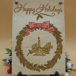 May include: Wooden sign with the words "Happy Holidays" in elegant script. The sign features a wreath design with a pink bow, holly, and a candle. The sign is held up by a black stand and has red string for hanging.