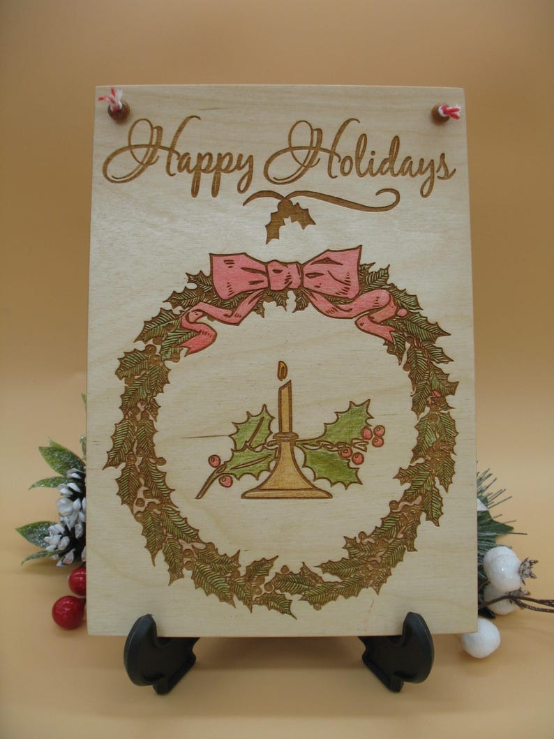 May include: Wooden sign with the words "Happy Holidays" in elegant script. The sign features a wreath design with a pink bow, holly, and a candle. The sign is held up by a black stand and has red string for hanging.