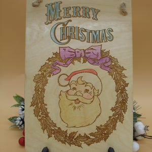 May include: Wooden Christmas sign featuring a Santa Claus illustration within a wreath, with the words "Merry Christmas" in a decorative font. The sign has a light wood grain background and a pink bow.