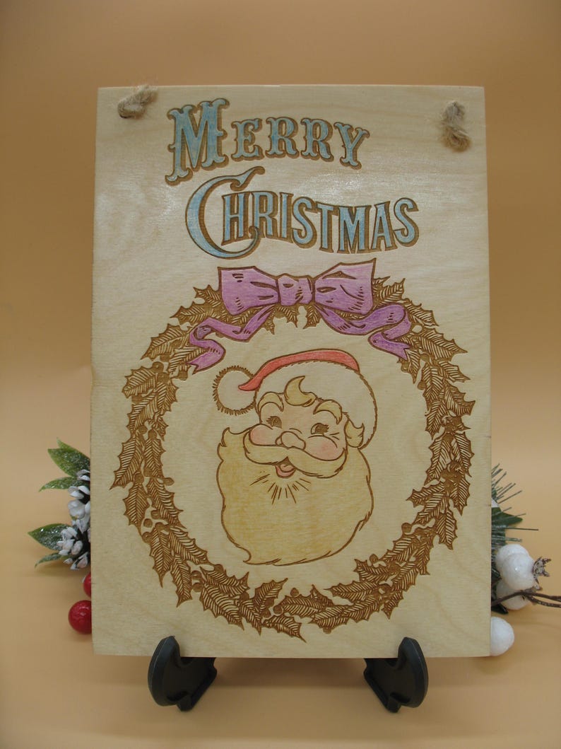 May include: Wooden Christmas sign featuring a Santa Claus illustration within a wreath, with the words "Merry Christmas" in a decorative font. The sign has a light wood grain background and a pink bow.