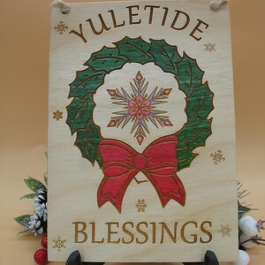 May include: Wooden sign with the words "Yuletide Blessings" and a wreath design. The wreath is green with a red bow and a snowflake in the center. The sign is light brown with a rope for hanging.