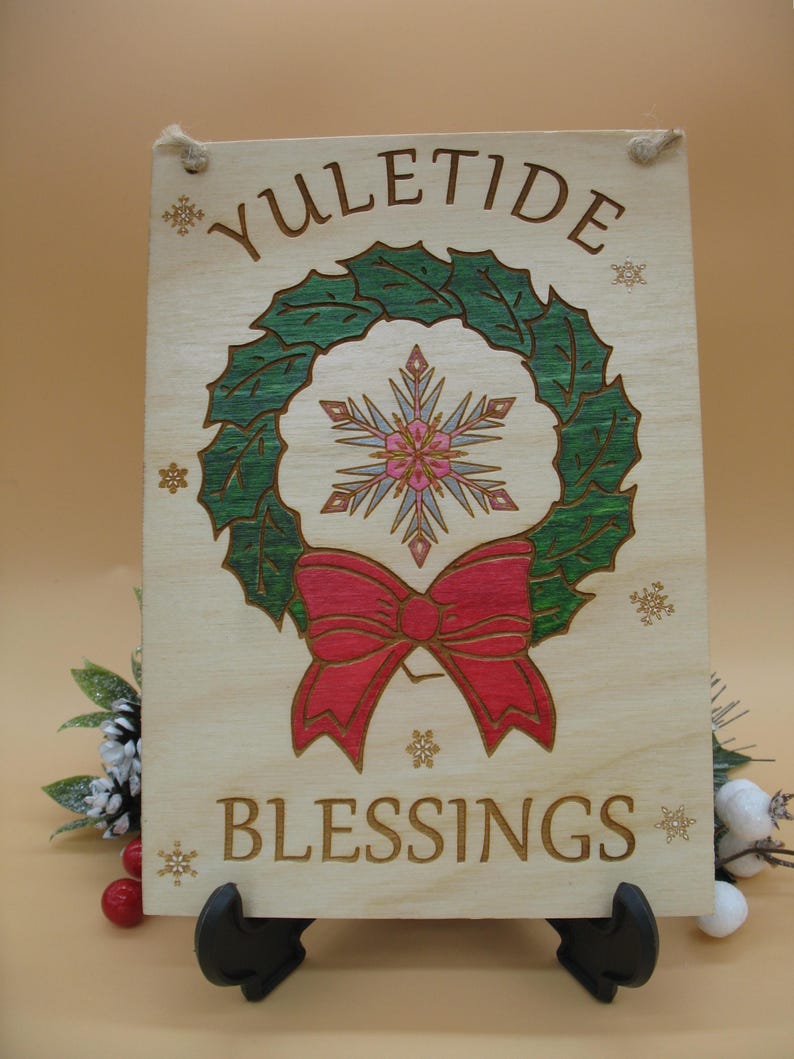 May include: Wooden sign with the words "Yuletide Blessings" and a wreath design. The wreath is green with a red bow and a snowflake in the center. The sign is light brown with a rope for hanging.