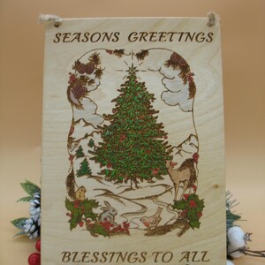 May include: Wooden Christmas sign with the words "SEASONS GREETINGS" and "BLESSINGS TO ALL". The sign features a detailed illustration of a Christmas tree, deer, rabbit, and other festive elements. The sign is held up by a black stand.