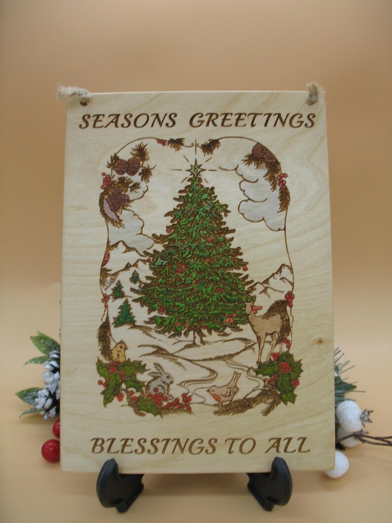 May include: Wooden Christmas sign with the words "SEASONS GREETINGS" and "BLESSINGS TO ALL". The sign features a detailed illustration of a Christmas tree, deer, rabbit, and other festive elements. The sign is held up by a black stand.