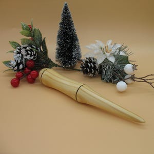 May include: A light-coloured wooden wand with a tapered design, set against a festive backdrop of miniature Christmas trees, pinecones, and red berries. The wand is the focal point, suggesting a theme of magic or holiday decor.