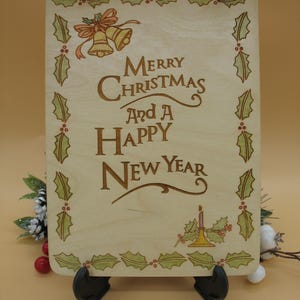 May include: Wooden Christmas sign with the message "Merry Christmas and a Happy New Year" in brown lettering. The sign is decorated with holly leaves, berries, and bells. A jute rope is attached for hanging.